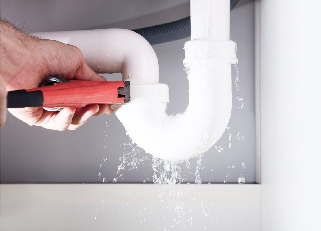 Hand hält roten Rohrschlüssel an weißen Siphon mit spritzendem Wasser unter Spüle.